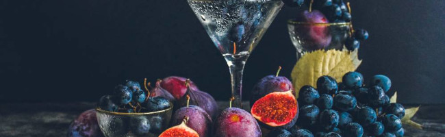 A collection of fruits, blueberries and bright figs, lay around a dewy wine glass upon a fine tablecloth. The atmosphere is dark and moody.